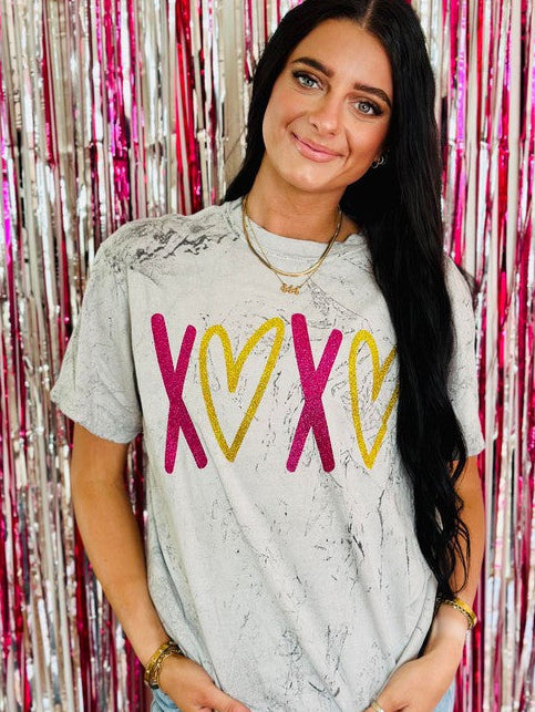 A person wearing a gray t-shirt with hot pink and gold glitter hearts design, standing in front of a pink glittery background.