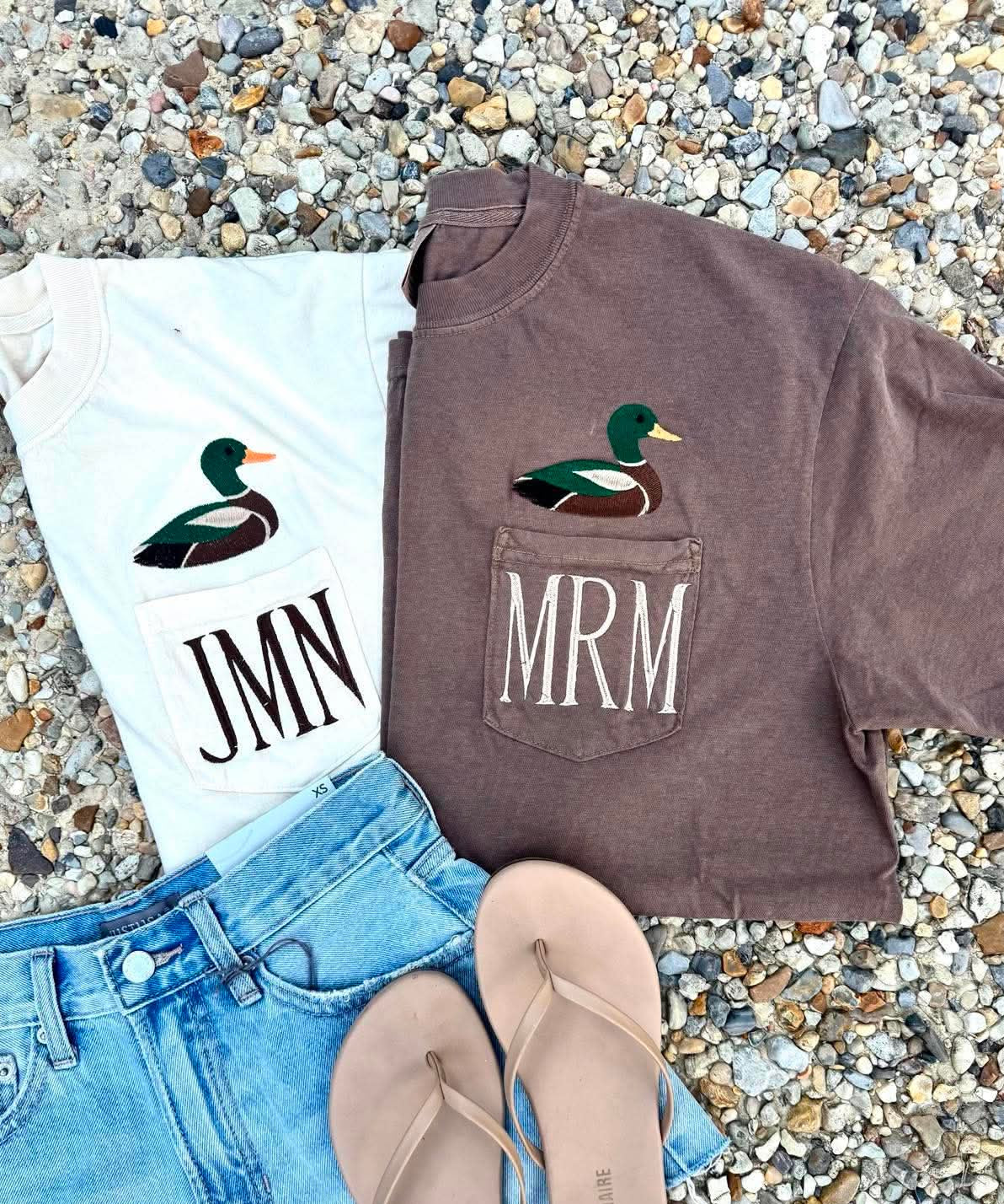 A styled flat-lay of two graphic t-shirts with duck illustrations and pocket monograms, paired with light blue denim shorts and beige flip-flops on a bed of small gray pebbles. One white t-shirt has a mallard graphic above the text 'JMN', and one brown t-shirt has a mallard graphic above the text 'MRM'.