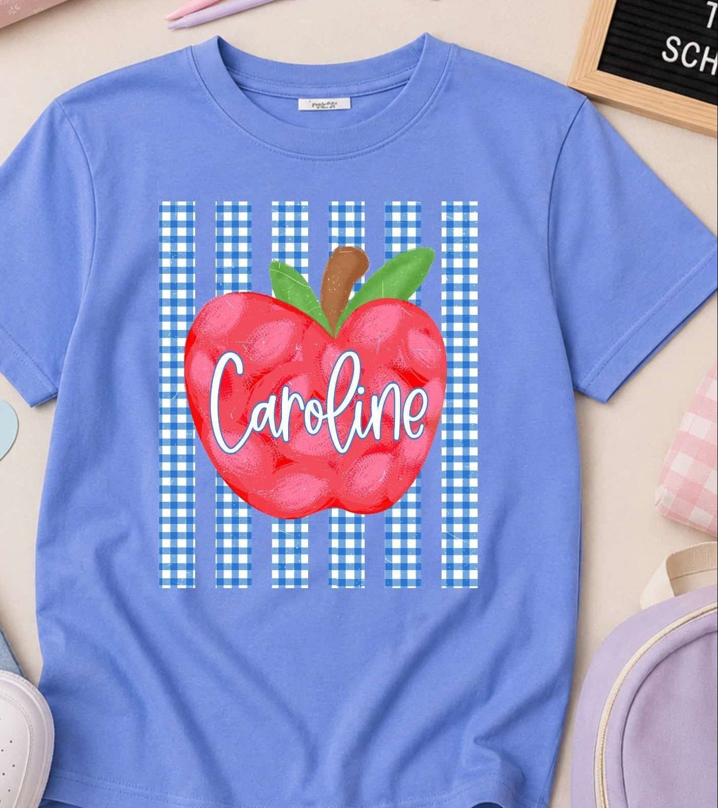 A personalized blue short-sleeve t-shirt with a red apple graphic and the name 'Caroline' printed on the front, styled in a flat lay with school accessories.