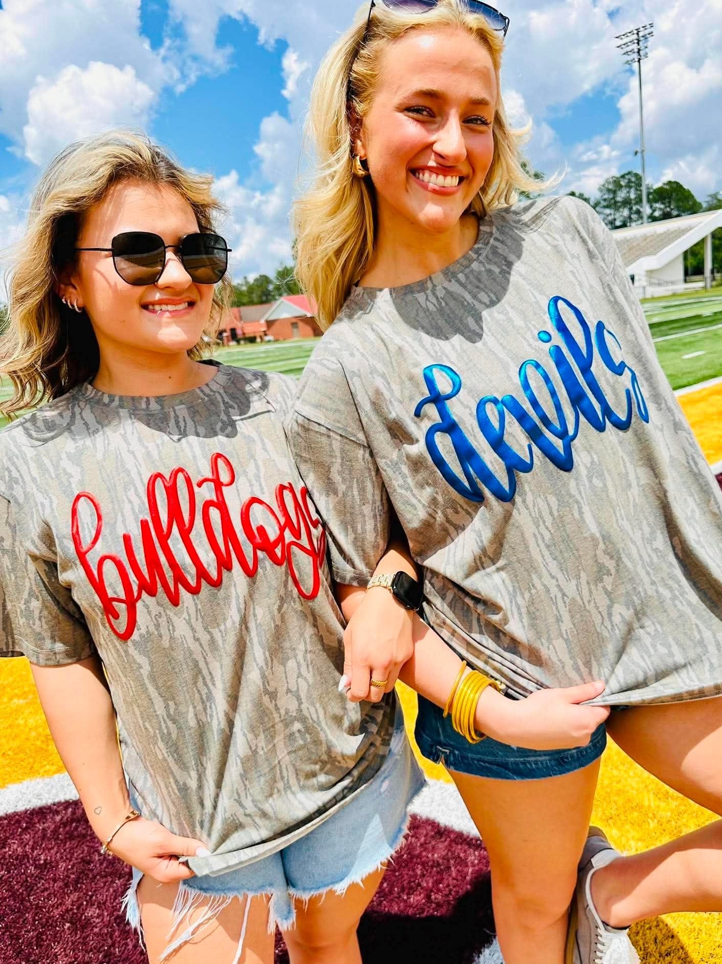 Two women wearing 'Bulldogs' and 'Devils' metallic puff shirts on a sports field.