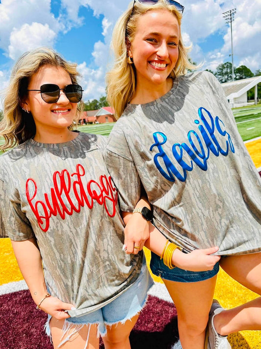 Two women wearing 'Bulldogs' and 'Devils' metallic puff shirts on a sports field.