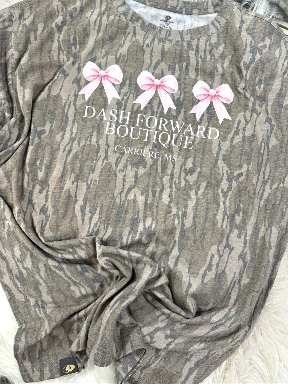 Mossy Oak Camouflage shirt with 'Dash Forward Boutique' text and pink bows on a white surface