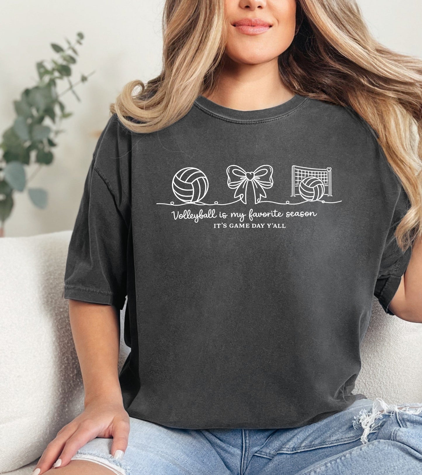 Person wearing a dark gray t-shirt with sports-themed graphics and Person wearing a dark gray t-shirt with a "Volleyball is my favorite season" graphic design and text, sitting on a couch. text.