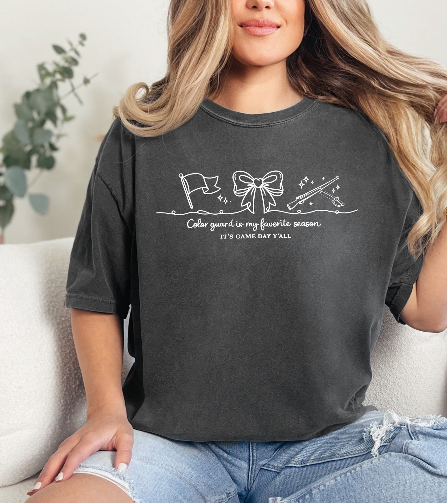 Person wearing a dark gray t-shirt with a "Color Guard is my favorite season" graphic design and text, sitting on a couch.