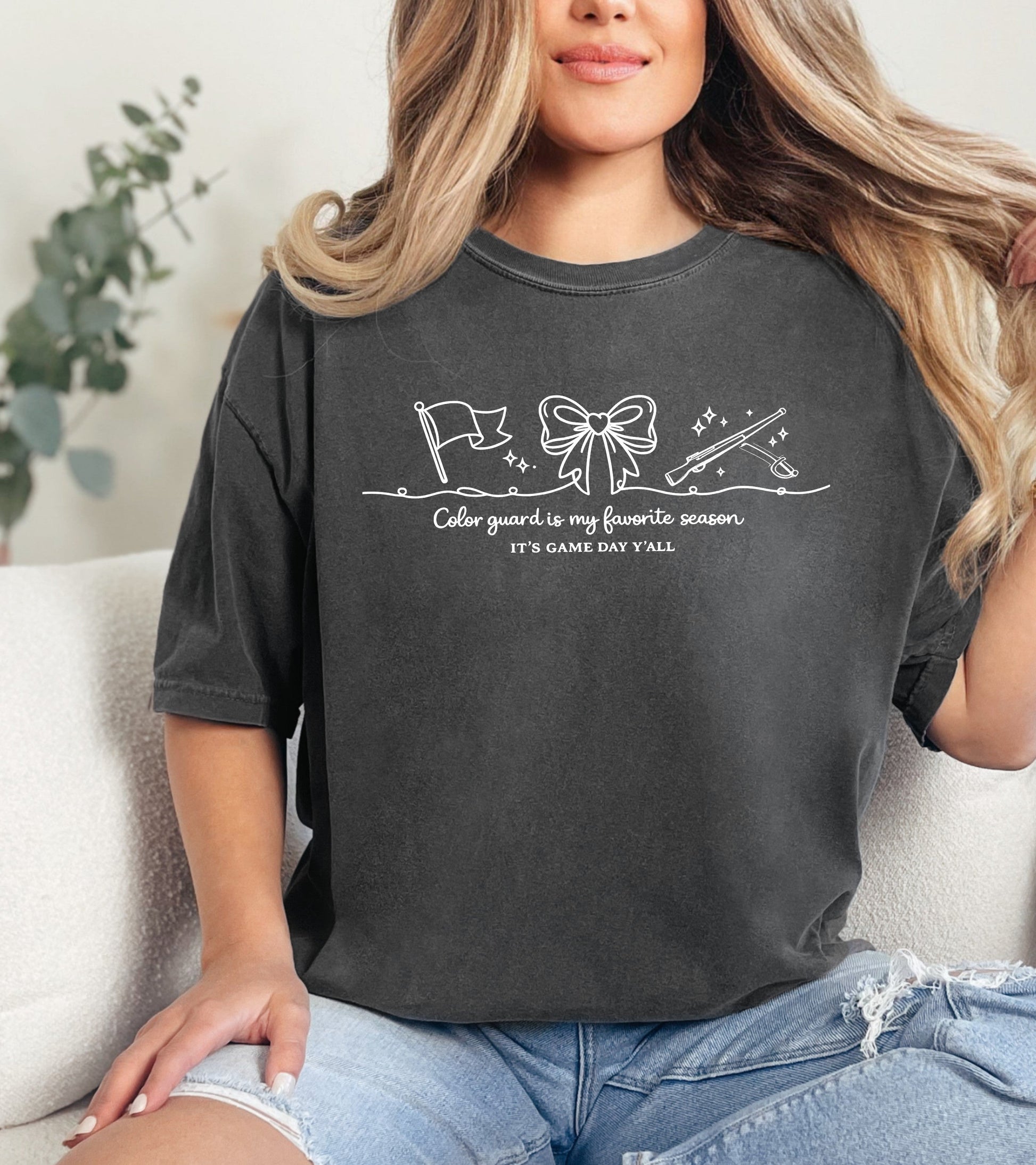 Person wearing a dark gray t-shirt with a "Color Guard is my favorite season" graphic design and text, sitting on a couch.