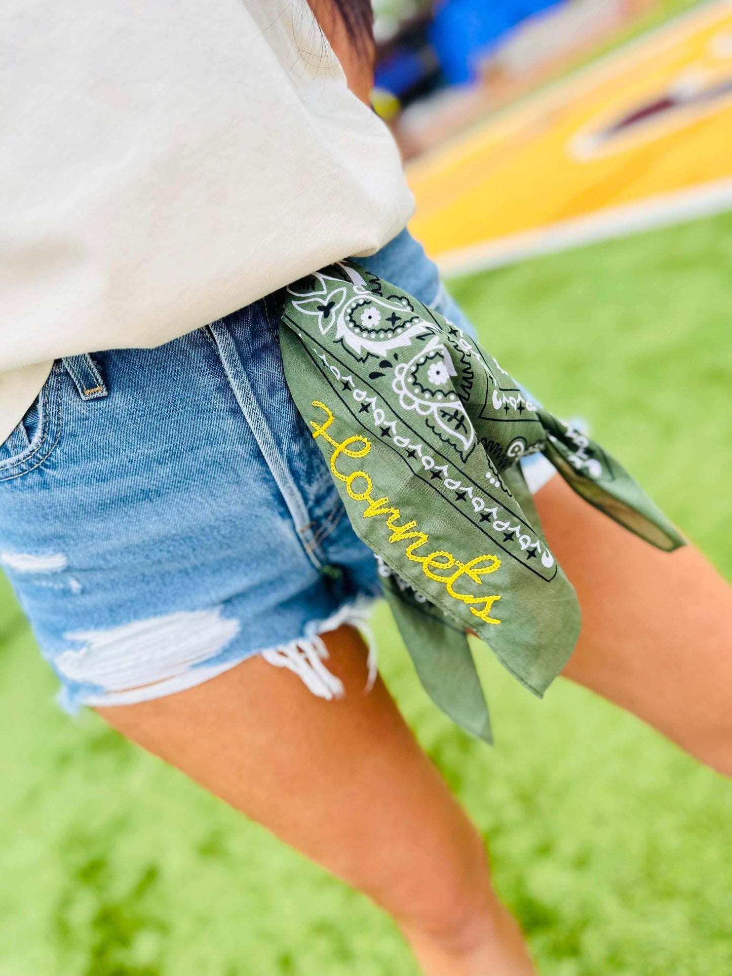 Person wearing denim shorts with a green bandana around their waist, featuring the word 'Hornets'.