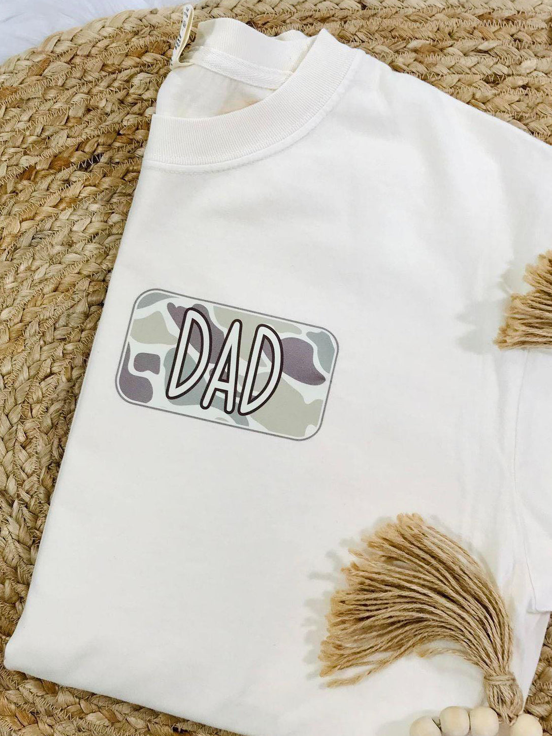 A white t-shirt with camouflage graphics and the text 'DAD' on the front pocket, displayed on a woven surface with beaded decorations in the background.