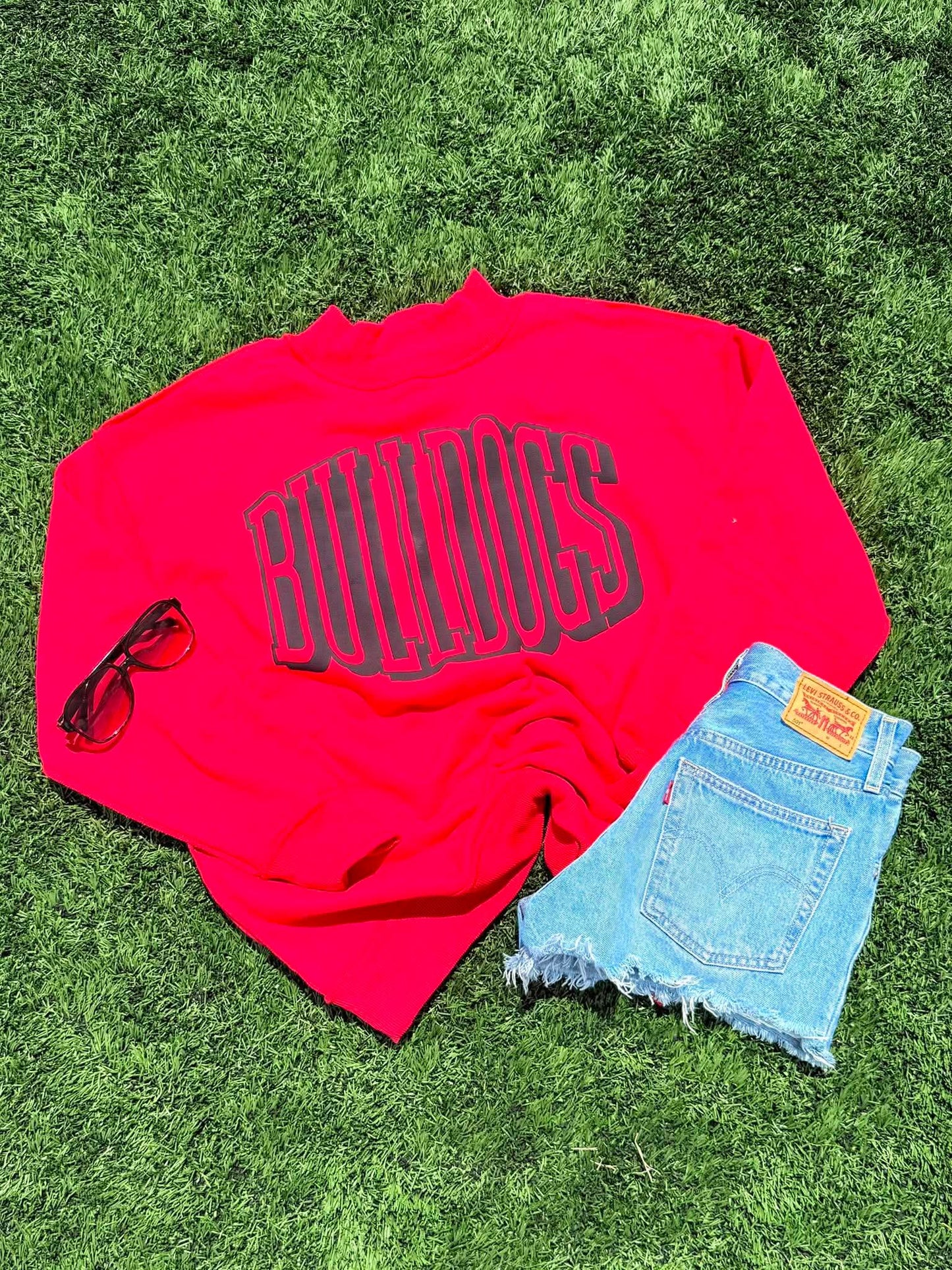 Red shirt with 'Bulldogs' text, sunglasses, and denim shorts on grass