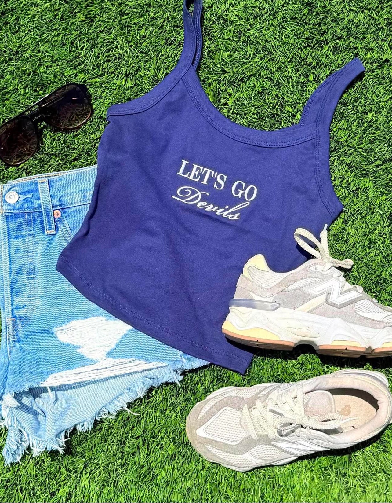 Blue tank top with 'LET'S GO' text, sunglasses, denim jacket, and sneakers on grass