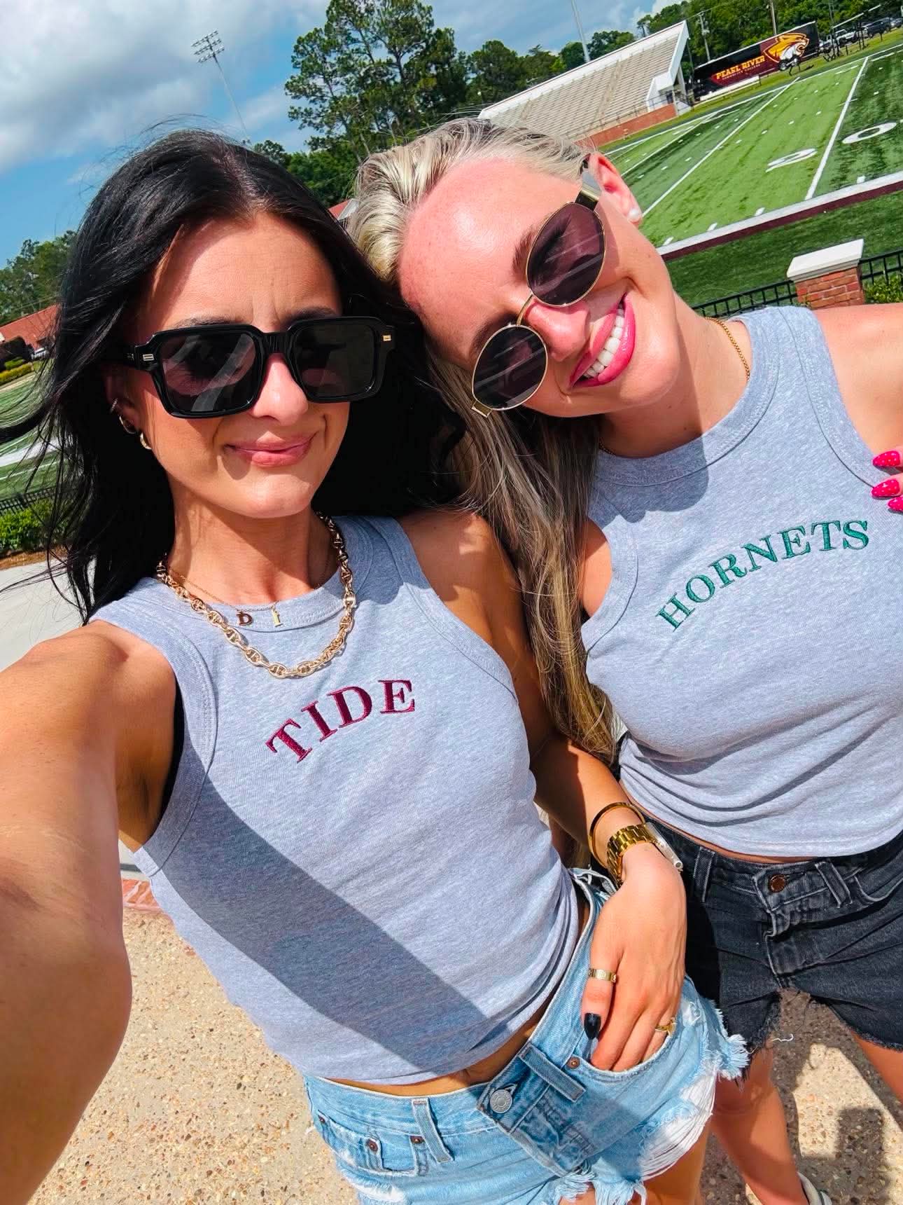 Two women wearing gray tank tops with 'TIDE' and 'Hornets' text, standing outdoors near a sports field.