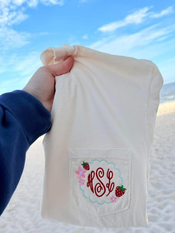 Beige fabric with a monogram and strawberry design held by a hand against a beach background