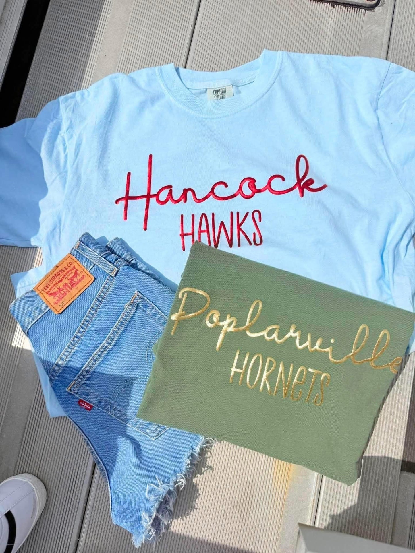 Light blue t-shirt with 'Hancock Hawks' text, Levi's jeans, and green tote bag with 'Poplarville Hornets' text on a wooden surface.