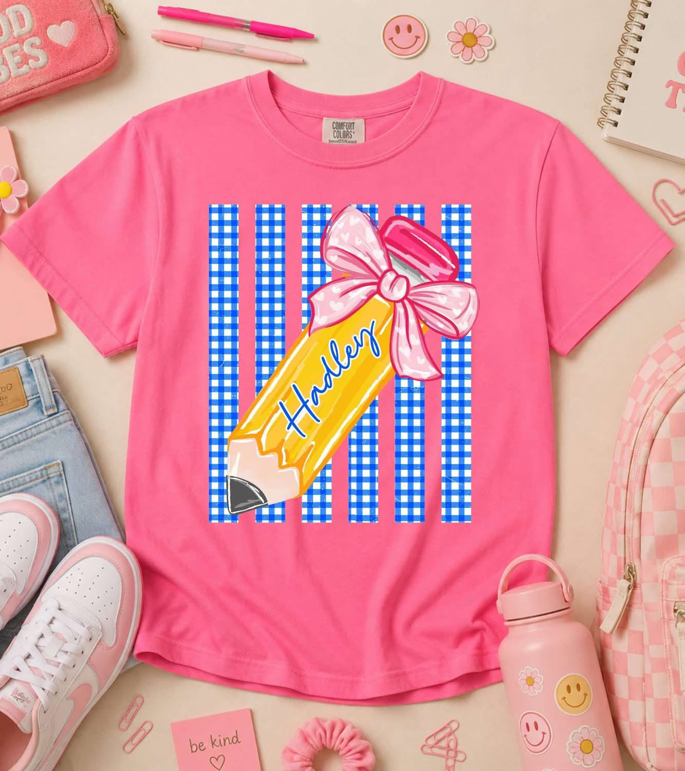 Pink t-shirt with a pencil graphic and 'Haley' text, surrounded by stationery items on a light background.