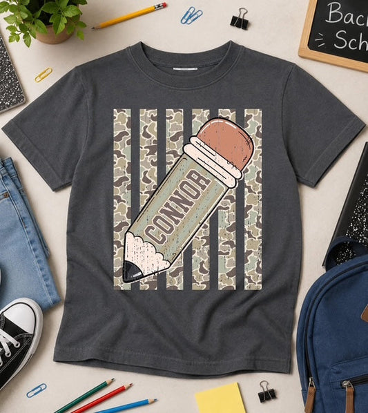 Gray t-shirt with a pencil graphic and 'CONNOR' text, surrounded by school supplies on a light surface.