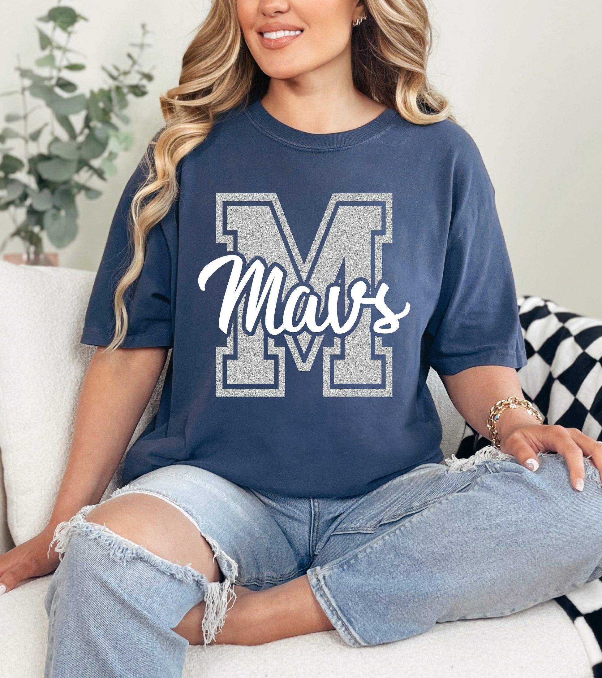 Woman wearing a navy blue t-shirt with 'Mavs' printed on it, sitting on a couch.