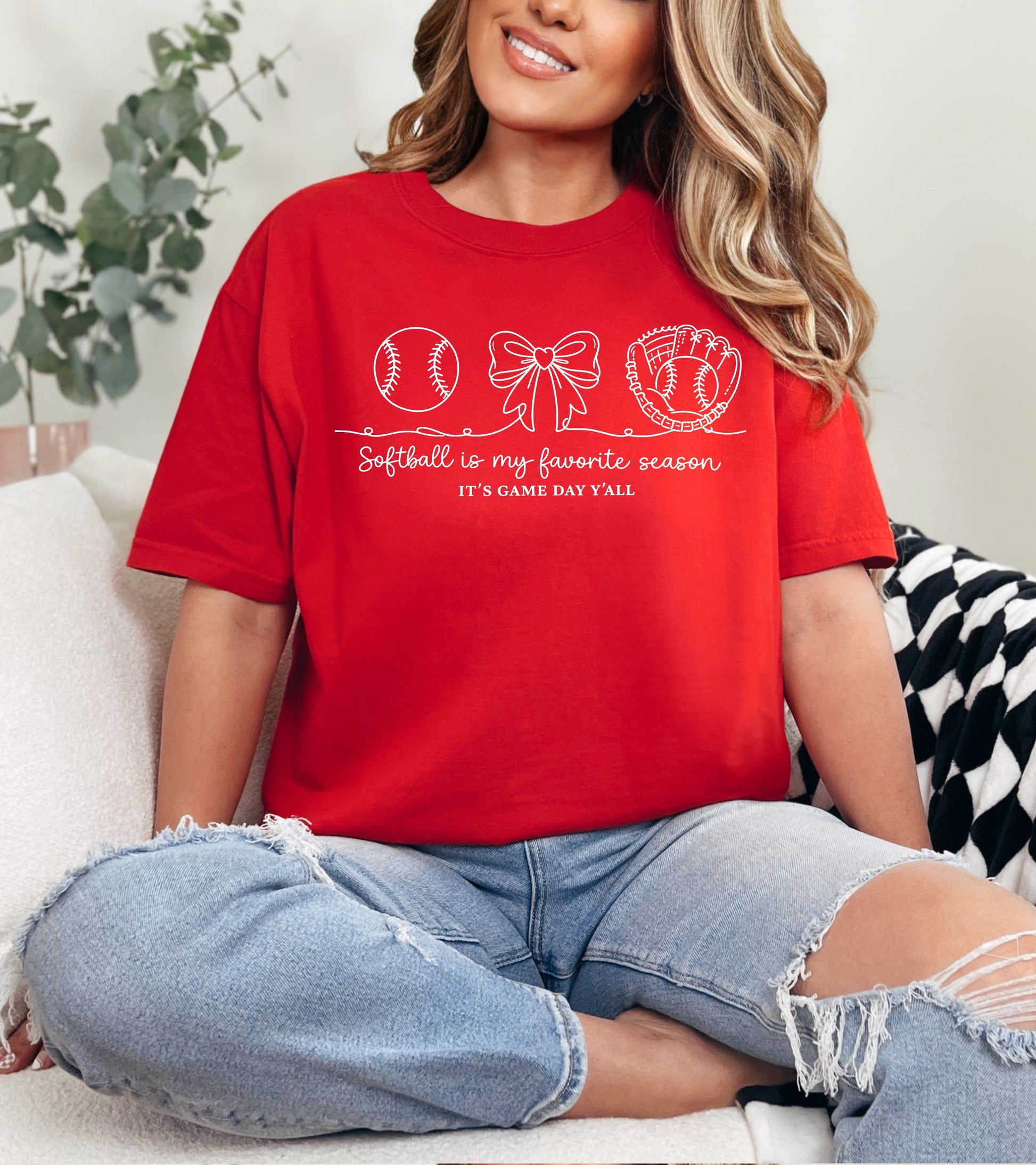 Person wearing a red t-shirt with sports-themed design and "Person wearing a dark gray t-shirt with a "Softball is my favorite season" graphic design and text, sitting on a couch" text, sitting on a couch.